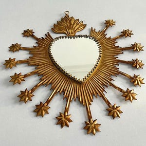 May include: A heart-shaped mirror with a gold frame and starburst design. The mirror has a decorative gold border and is surrounded by radiating gold rays and star-shaped accents. A decorative gold element is at the top.