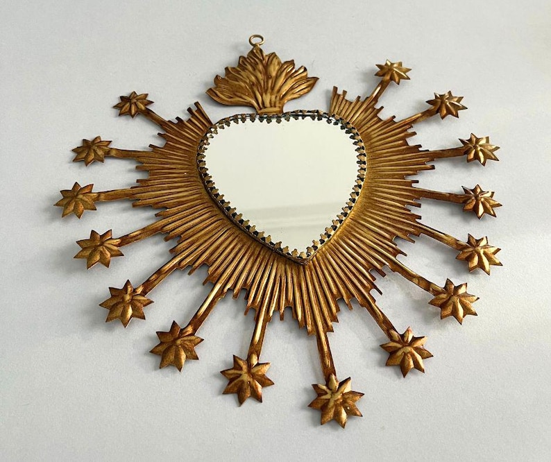 May include: A heart-shaped mirror with a gold frame and starburst design. The mirror has a decorative gold border and is surrounded by radiating gold rays and star-shaped accents. A decorative gold element is at the top.