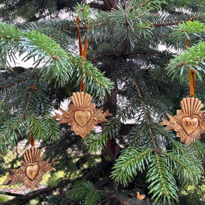 May include: Ornate, gold-colored Christmas ornaments hanging on a green pine tree. Each ornament features a heart design with the word "Love" inscribed. The ornaments are suspended by orange ribbons, creating a festive holiday display.