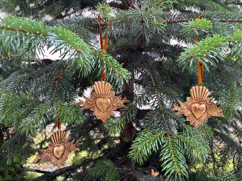 May include: Ornate, gold-colored Christmas ornaments hanging on a green pine tree. Each ornament features a heart design with the word "Love" inscribed. The ornaments are suspended by orange ribbons, creating a festive holiday display.