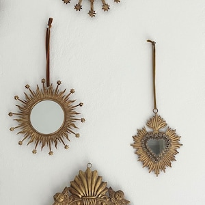 May include: A collection of three decorative, gold-colored mirrors with heart and sunburst designs. The mirrors are hanging on a white wall, each with a different shape and size, and are suitable for home decor.
