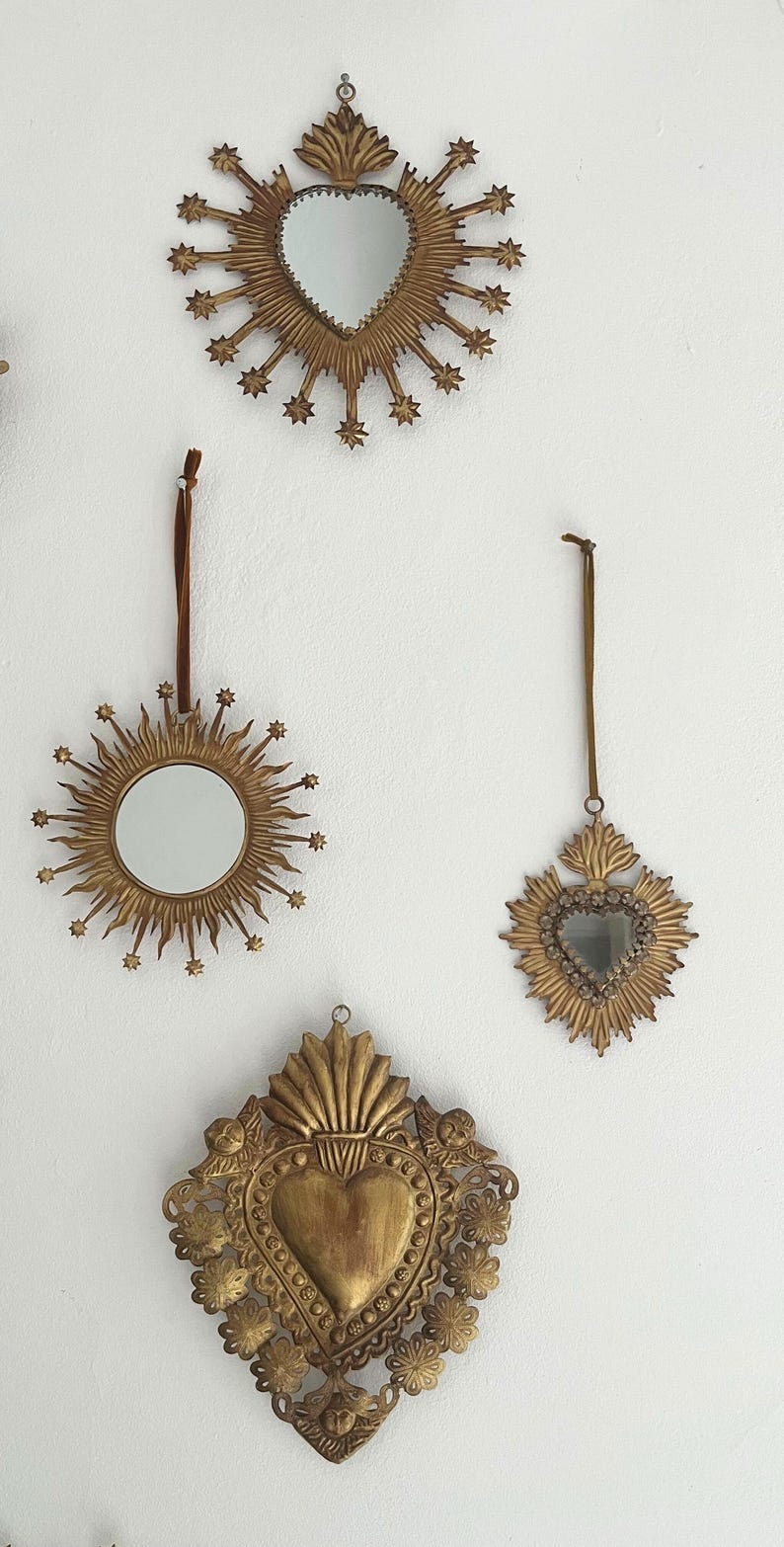 May include: A collection of three decorative, gold-colored mirrors with heart and sunburst designs. The mirrors are hanging on a white wall, each with a different shape and size, and are suitable for home decor.