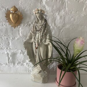 May include: A white plaster statue of the Virgin Mary wearing a flower crown and a beaded necklace. The statue is standing on a white surface in front of a white brick wall. A gold heart with flames is on the wall behind the statue. A pink potted plant is on the right side of the image.