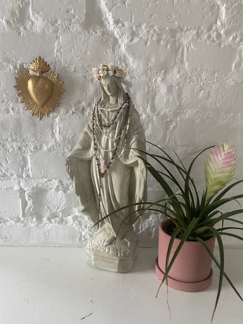 May include: A white plaster statue of the Virgin Mary wearing a flower crown and a beaded necklace. The statue is standing on a white surface in front of a white brick wall. A gold heart with flames is on the wall behind the statue. A pink potted plant is on the right side of the image.