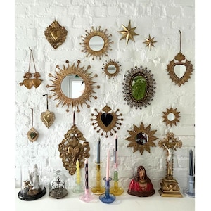 May include: A collection of gold-toned decorative mirrors, stars, and heart-shaped ornaments hang on a white brick wall. Below, a shelf displays candles, a religious statue, and a crucifix. The mirrors vary in size and shape, creating a vintage aesthetic.
