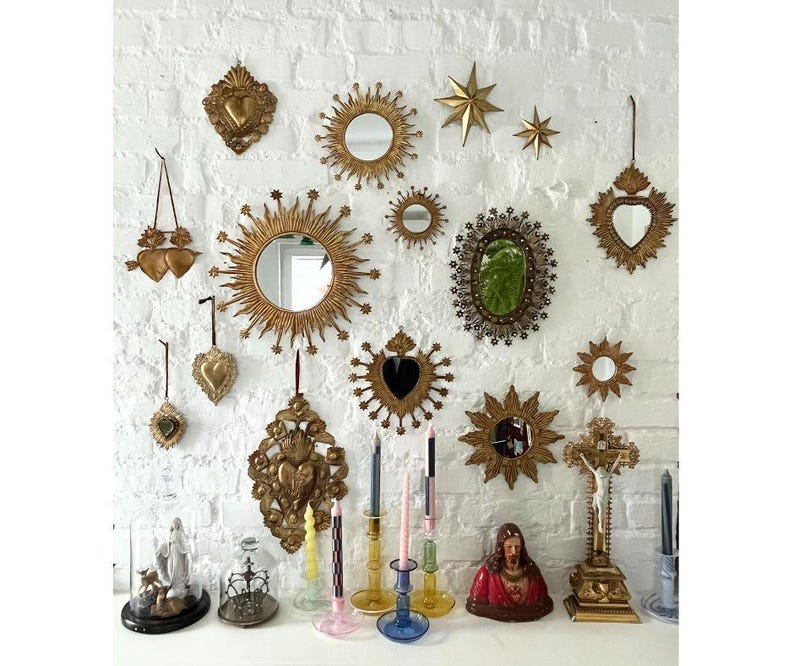 May include: A collection of gold-toned decorative mirrors, stars, and heart-shaped ornaments hang on a white brick wall. Below, a shelf displays candles, a religious statue, and a crucifix. The mirrors vary in size and shape, creating a vintage aesthetic.