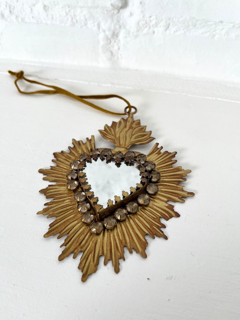 May include: A heart-shaped mirror with a gold-colored frame and a sunburst design. The mirror is surrounded by small, clear gemstones. A brown cord is attached for hanging. The item is likely a decorative wall hanging.