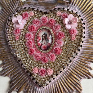 Puede incluir: Un icono religioso en forma de coraz&oacute;n con una imagen central de un santo rodeado de rosas rosas. El coraz&oacute;n est&aacute; adornado con pedrer&iacute;a y detalles florales rosas, sobre un fondo dorado y rosa.