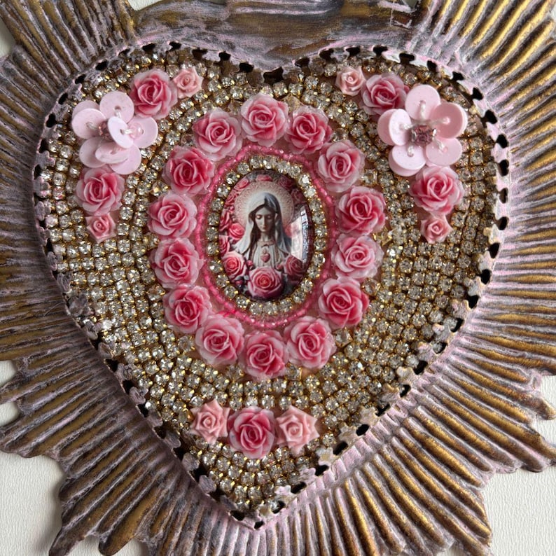 Puede incluir: Un icono religioso en forma de coraz&oacute;n con una imagen central de un santo rodeado de rosas rosas. El coraz&oacute;n est&aacute; adornado con pedrer&iacute;a y detalles florales rosas, sobre un fondo dorado y rosa.