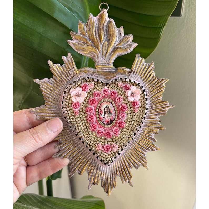 Puede incluir: Adorno de pared en forma de coraz&oacute;n con acabado dorado y rosa. El coraz&oacute;n presenta una imagen central de una figura religiosa, rodeada de rosas rosas y adornos brillantes. El dise&ntilde;o incluye un patr&oacute;n de rayos de sol.