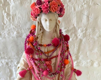May include: A white bust statue adorned with a vibrant floral crown featuring shades of pink, coral, and red. The figure is draped in multiple layered necklaces consisting of pink and orange beads, metallic chains, and small fabric pom-poms.