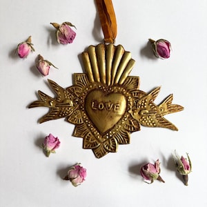 May include: A gold-colored metal heart-shaped ornament with the word "LOVE" in the center, surrounded by decorative elements and two bird figures. The ornament hangs from a brown ribbon and is accompanied by several dried pink rose buds.