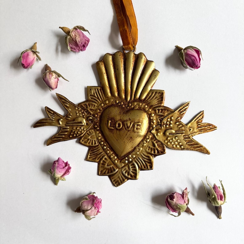 May include: A gold-colored metal heart-shaped ornament with the word "LOVE" in the center, surrounded by decorative elements and two bird figures. The ornament hangs from a brown ribbon and is accompanied by several dried pink rose buds.
