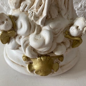 May include: White ceramic sculpture of a woman with two cherubs, one on each side. The woman is wearing a flowing robe and the cherubs have wings. The sculpture has gold accents.