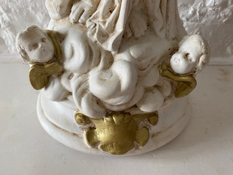 May include: White ceramic sculpture of a woman with two cherubs, one on each side. The woman is wearing a flowing robe and the cherubs have wings. The sculpture has gold accents.