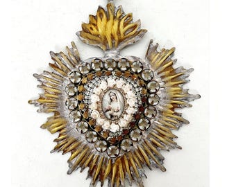 May include: A decorative heart-shaped wall hanging featuring a central oval portrait of a figure in a veil, surrounded by small white floral accents and clear rhinestones. The frame has a distressed gold and silver finish with radiating rays.