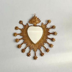May include: A heart-shaped mirror with a gold-colored frame. The frame features a sunburst design with star-shaped accents. The mirror is hanging on a white wall.