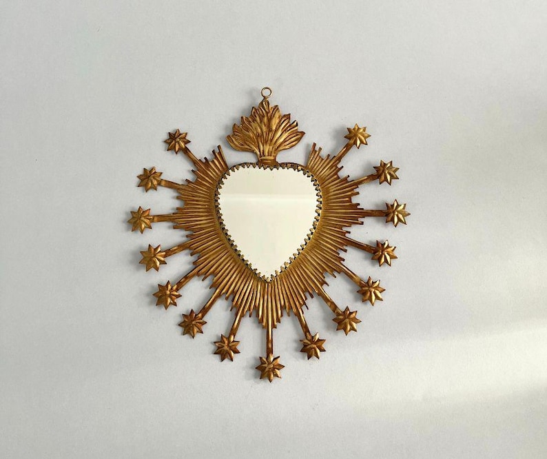 May include: A heart-shaped mirror with a gold-colored frame. The frame features a sunburst design with star-shaped accents. The mirror is hanging on a white wall.