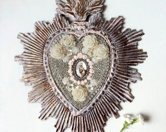 May include: A heart-shaped wall hanging with a distressed, metallic finish. The heart is decorated with white roses, clear beads, and a central religious image. The design includes a sunburst frame and floral accents.