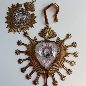 May include: Two ornate, gold-toned religious ornaments. One is a sunburst design with a central image and clear crystal accents. The other is heart-shaped, with a central image, pink roses, and angel figurines, surrounded by starburst rays.