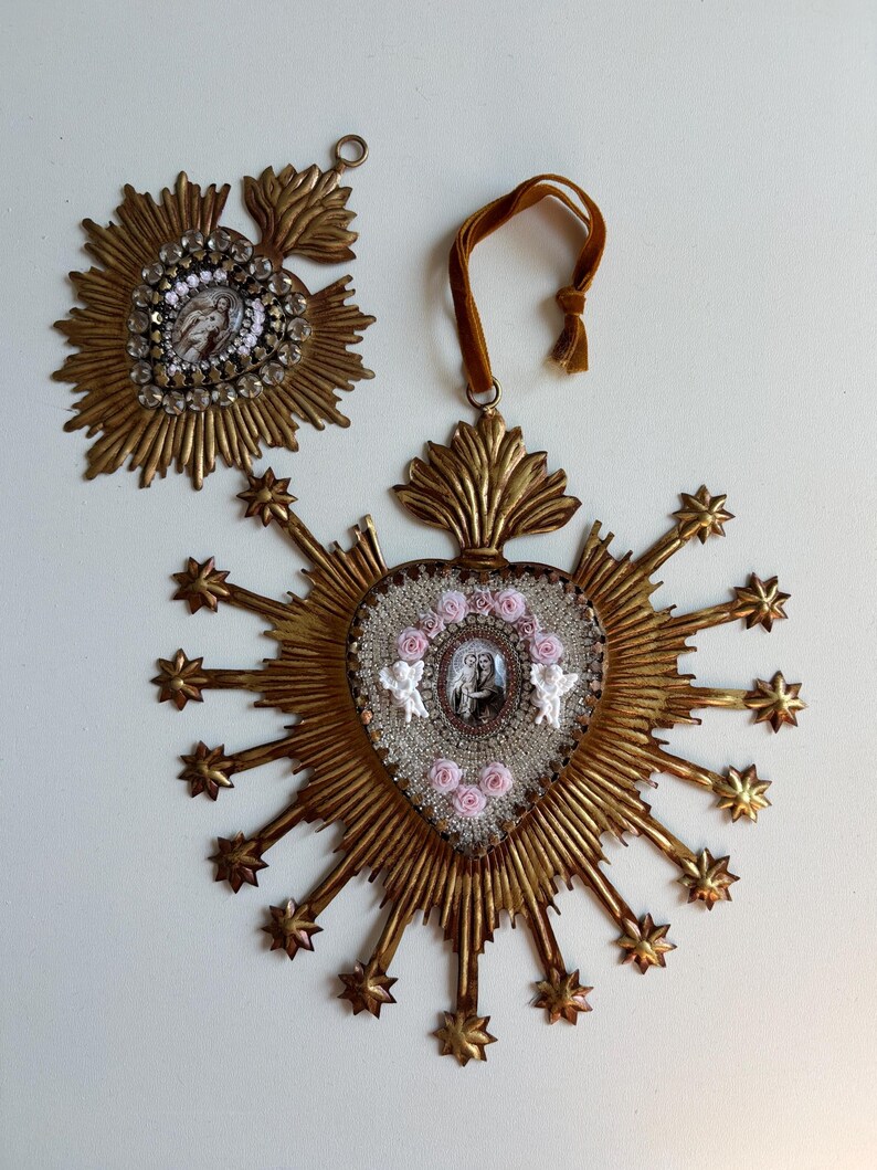 May include: Two ornate, gold-toned religious ornaments. One is a sunburst design with a central image and clear crystal accents. The other is heart-shaped, with a central image, pink roses, and angel figurines, surrounded by starburst rays.