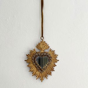 May include: A gold-colored, heart-shaped wall hanging with a sunburst design. The center features a heart-shaped mirror surrounded by small, clear stones. A brown cord is attached for hanging.