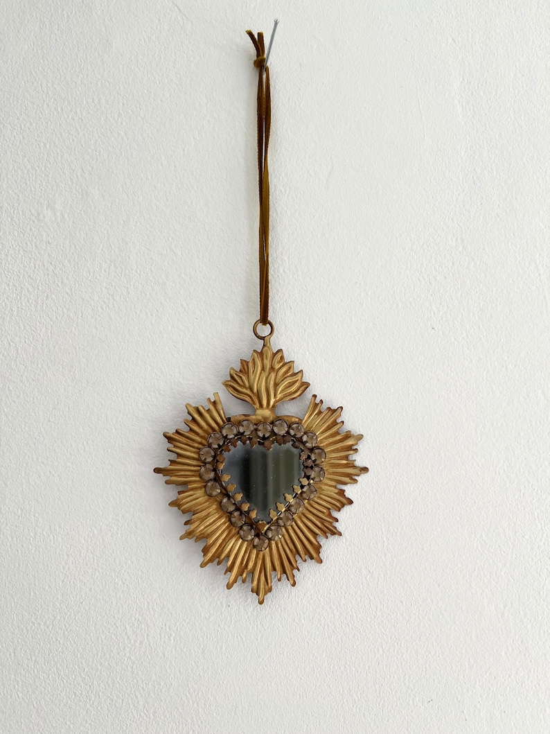 May include: A gold-colored, heart-shaped wall hanging with a sunburst design. The center features a heart-shaped mirror surrounded by small, clear stones. A brown cord is attached for hanging.