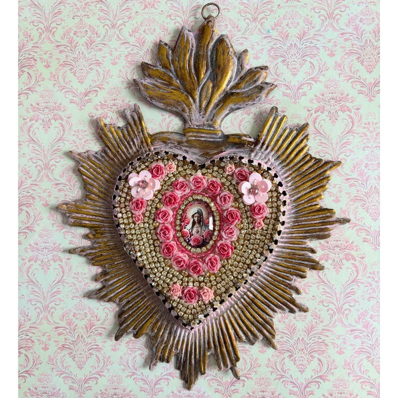 Puede incluir: Adorno de pared en forma de coraz&oacute;n dorado, con una imagen central de una santa rodeada de rosas rosas y detalles brillantes. El coraz&oacute;n est&aacute; enmarcado por un dise&ntilde;o de rayos de sol y coronado por un elemento decorativo.