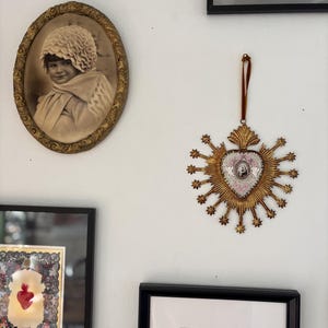 May include: A wall display featuring framed art and decorative pieces. An oval-framed sepia-toned portrait of a child hangs near a gold-colored, starburst-shaped ornament with a central image. Other framed artworks are also visible.