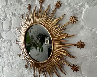 May include: A gold sunburst mirror with a round mirror in the centre. The mirror is surrounded by a decorative frame with a sunburst design and stars.