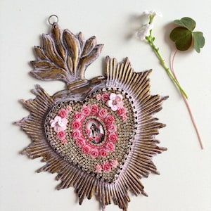 Puede incluir: Un adorno religioso en forma de coraz&oacute;n con una imagen central de un santo, rodeado de rosas rosas y pedrer&iacute;a. El adorno tiene un dise&ntilde;o de sol dorado y rosa y una parte superior decorativa. Una ramita de flores blancas y un tr&eacute;bol est&aacute;n junto al adorno.