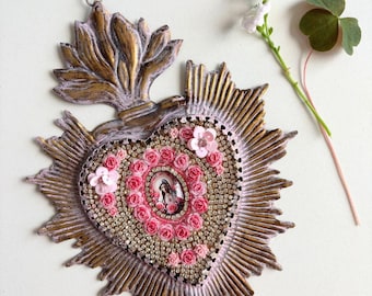 May include: A heart-shaped religious ornament featuring a central image of a saint, encircled by pink roses and rhinestones. The ornament has a gold and pink sunburst design and a decorative top. A sprig of white flowers and a clover are next to the ornament.