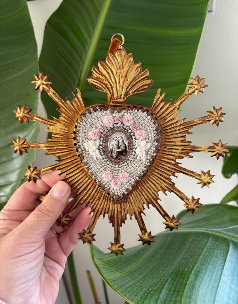 May include: A gold-colored, heart-shaped religious icon with a central image of a religious figure. The heart is surrounded by a sunburst design with star-shaped accents. The heart is decorated with pink roses and white beads.