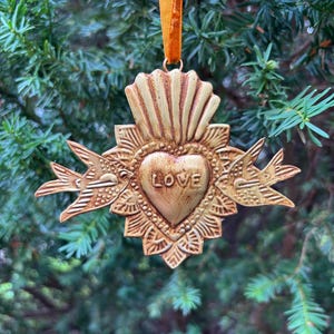 May include: A gold-colored heart-shaped ornament with the word "LOVE" in the center. The ornament has a textured surface and is adorned with decorative elements, including two birds on either side. It hangs from a gold ribbon, perfect for holiday decor.