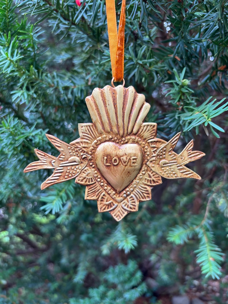May include: A gold-colored heart-shaped ornament with the word "LOVE" in the center. The ornament has a textured surface and is adorned with decorative elements, including two birds on either side. It hangs from a gold ribbon, perfect for holiday decor.