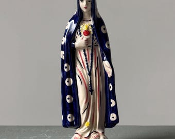 May include: A ceramic statue of a religious figure, with a blue cloak with white circles, a white dress with red and white stripes, and a heart-shaped object. The statue is on a cloud-like base.