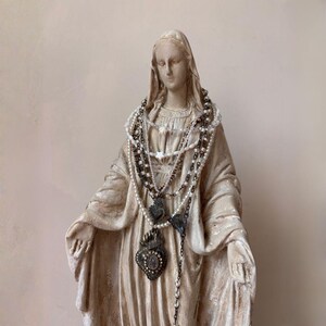 Puede incluir: Estatua de una figura religiosa, adornada con m&uacute;ltiples collares de cuentas y colgantes. La estatua es de color beige claro, y los collares incluyen perlas y adornos de metal.