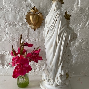 May include: White plaster statue of the Virgin Mary holding a baby Jesus. The statue is standing on a white base and has a gold crown. A gold heart with flames is on the wall behind the statue.