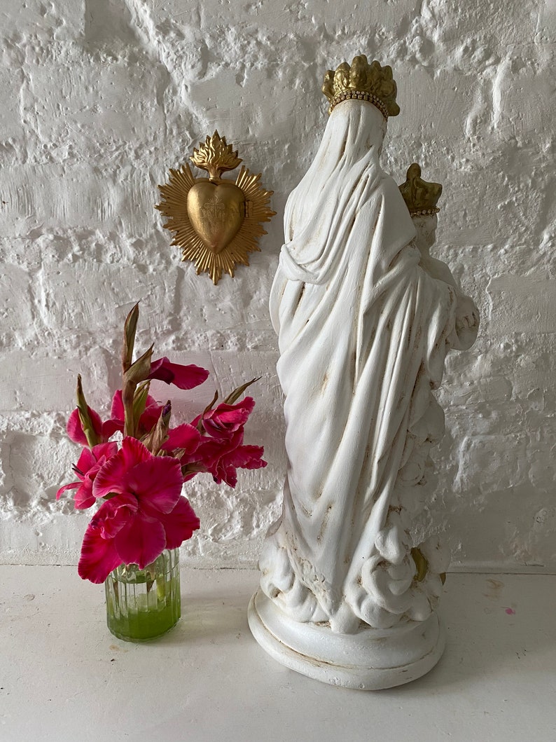 May include: White plaster statue of the Virgin Mary holding a baby Jesus. The statue is standing on a white base and has a gold crown. A gold heart with flames is on the wall behind the statue.