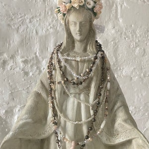 May include: A white plaster statue of the Virgin Mary wearing a crown of white and pink fabric flowers and multiple beaded necklaces.