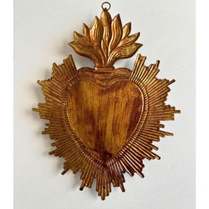 Puede incluir: Adorno de pared dorado en forma de coraz&oacute;n con dise&ntilde;o de rayos de sol. El coraz&oacute;n tiene una superficie texturizada y est&aacute; rematado con un elemento en forma de llama. La pieza est&aacute; dise&ntilde;ada para colgarse y tiene una est&eacute;tica r&uacute;stica y vintage.