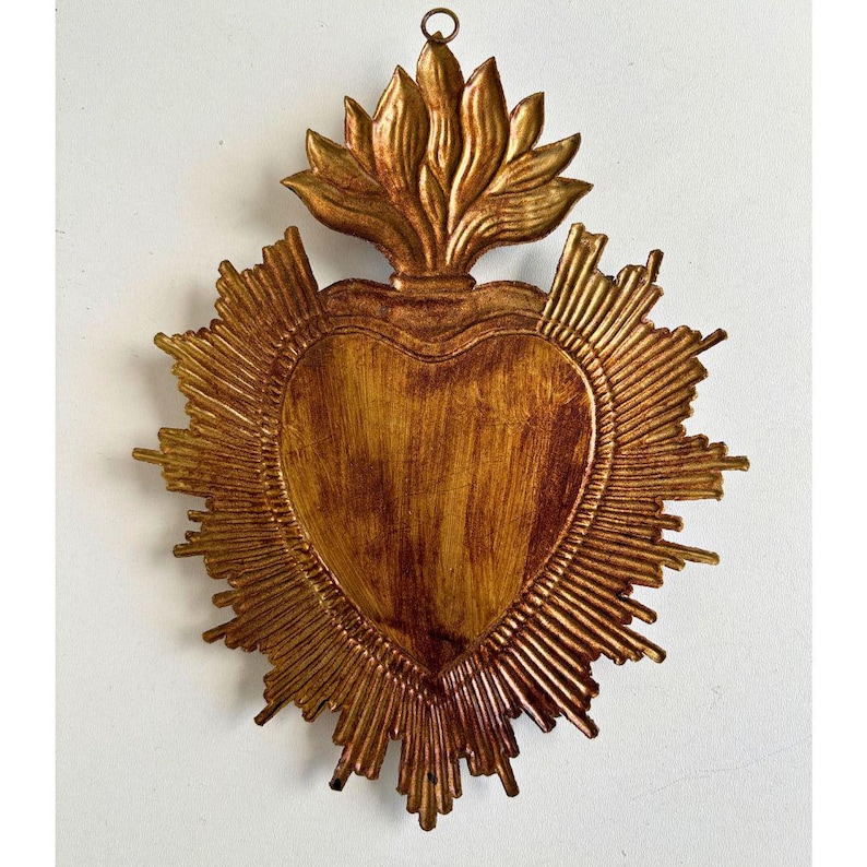 Puede incluir: Adorno de pared dorado en forma de coraz&oacute;n con dise&ntilde;o de rayos de sol. El coraz&oacute;n tiene una superficie texturizada y est&aacute; rematado con un elemento en forma de llama. La pieza est&aacute; dise&ntilde;ada para colgarse y tiene una est&eacute;tica r&uacute;stica y vintage.
