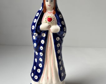 May include: A ceramic figurine of a religious figure, with a blue cloak dotted with white spots over a white dress. The figure has red hair and holds a heart with rays. The base is decorated with blue and white circles.