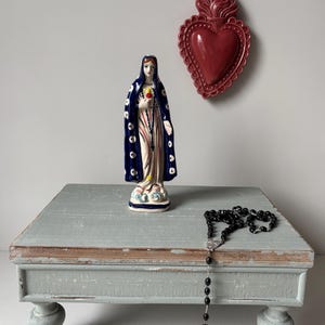 Ceramic Virgin Mary figurine displayed on wooden altar table with rosary and sacred heart wall decoration