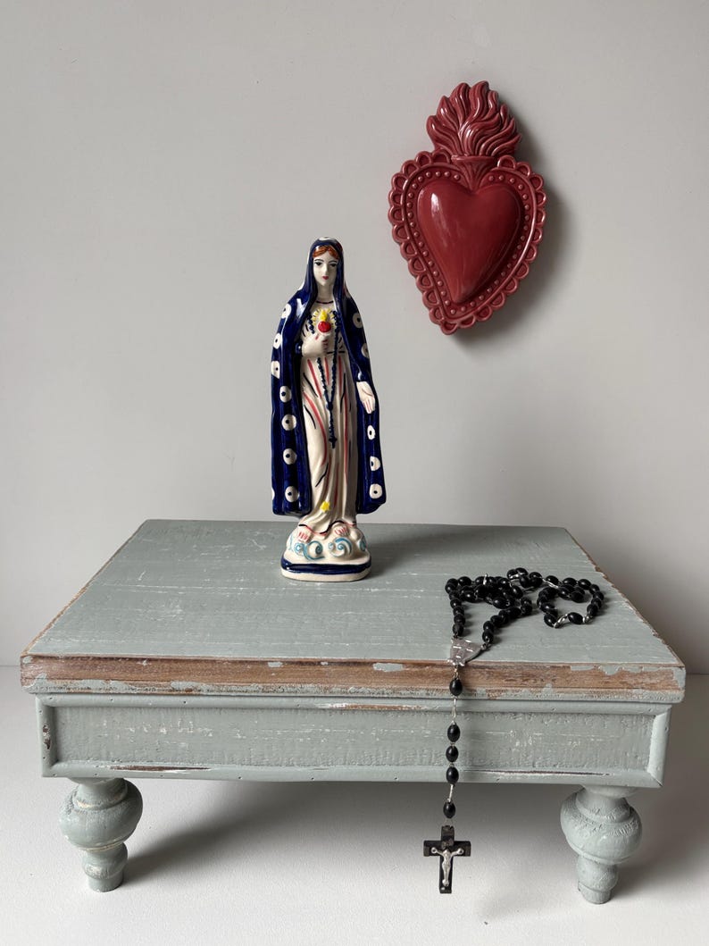 Ceramic Virgin Mary figurine displayed on wooden altar table with rosary and sacred heart wall decoration