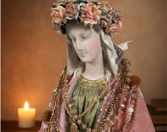 May include: A statue of a woman adorned with a floral crown and multiple beaded necklaces. The statue is painted in soft hues, featuring a pink robe and a gold dress. A lit candle is visible in the background.