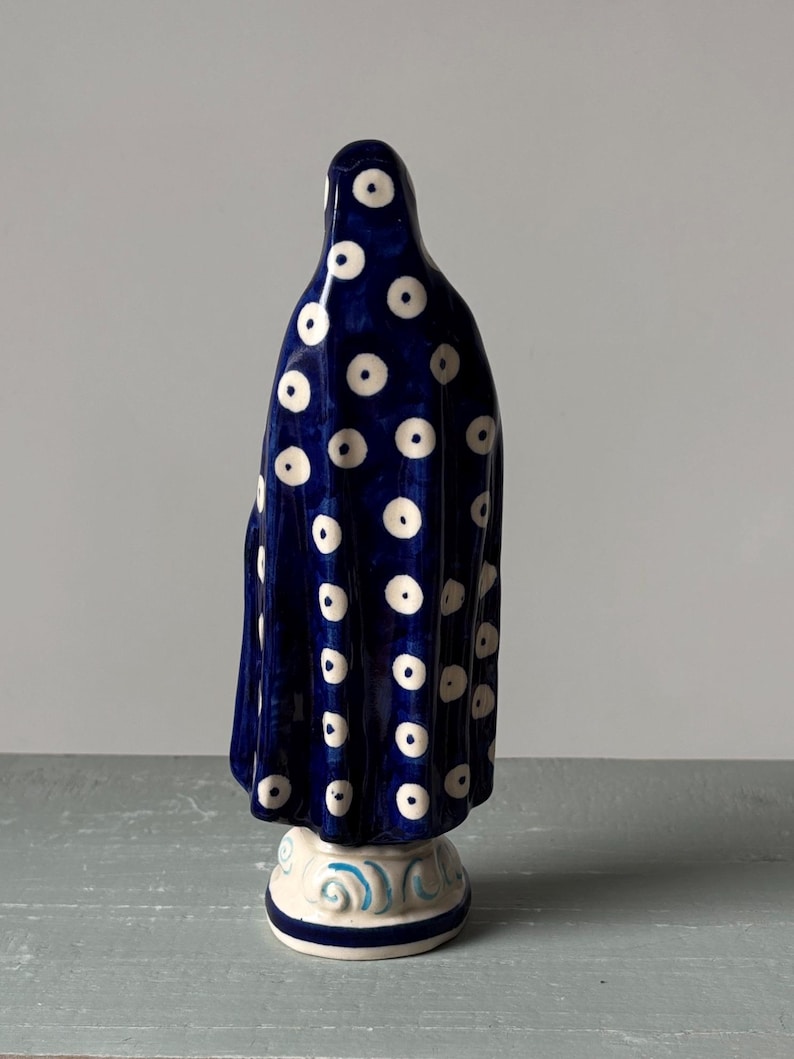 May include: A ceramic figurine of a draped figure, viewed from the back. The figure is primarily dark blue with a pattern of white polka dots. The base is white with blue decorative swirls.