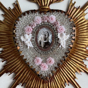 May include: A gold-colored, heart-shaped religious icon with a central oval image of Mary and Jesus. The heart is surrounded by clear beads, pink roses, and two white cherubs. The icon has a sunburst design.