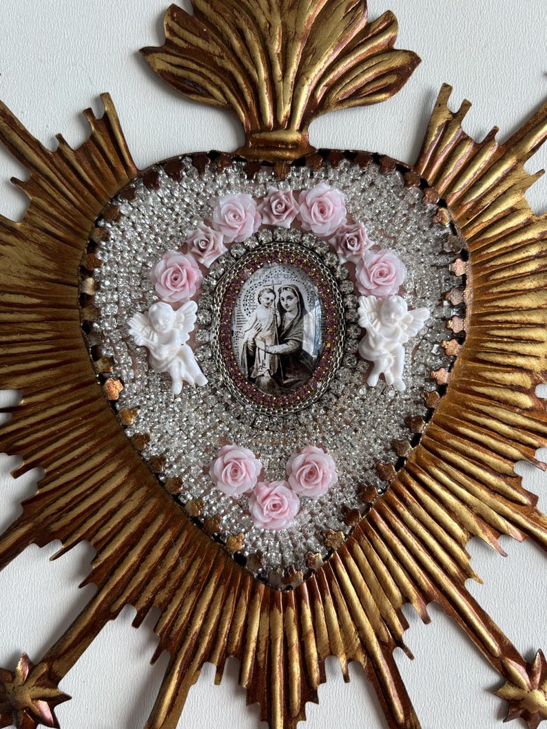 May include: A gold-colored, heart-shaped religious icon with a central oval image of Mary and Jesus. The heart is surrounded by clear beads, pink roses, and two white cherubs. The icon has a sunburst design.