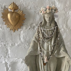 May include: A white statue of the Virgin Mary wearing a crown of white and pink flowers. She is adorned with multiple beaded necklaces, including a rosary with a tassel. The statue is set against a white brick wall with a gold heart-shaped religious icon hanging above.